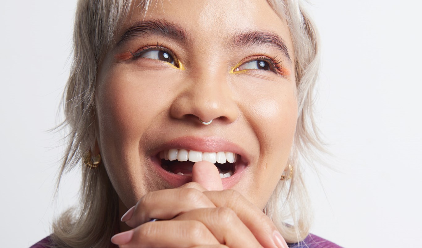 Kvinne med sminke, nesepiercing og kort, blondt hår, som smiler med åpen munn, ser til siden og har hendene knyttet foran haken mot en hvit bakgrunn.